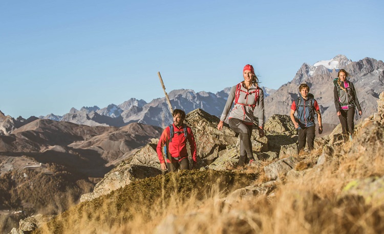 Vier wandelaars met rugzakken wandelen over een rotsachtig bergpad in een zonnig hooggebergte, met een achtergrond van indrukwekkende bergtoppen en een blauwe hemel.